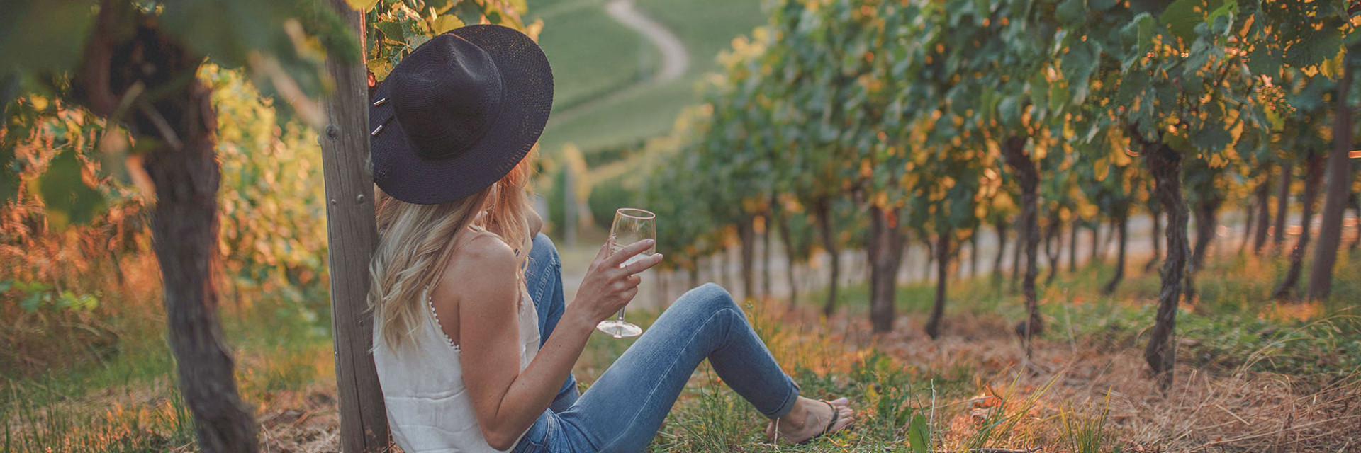 Foto einer Frau, die bei Sonnenuntergang über den Weinreben ein Glas Weißwein genießt.