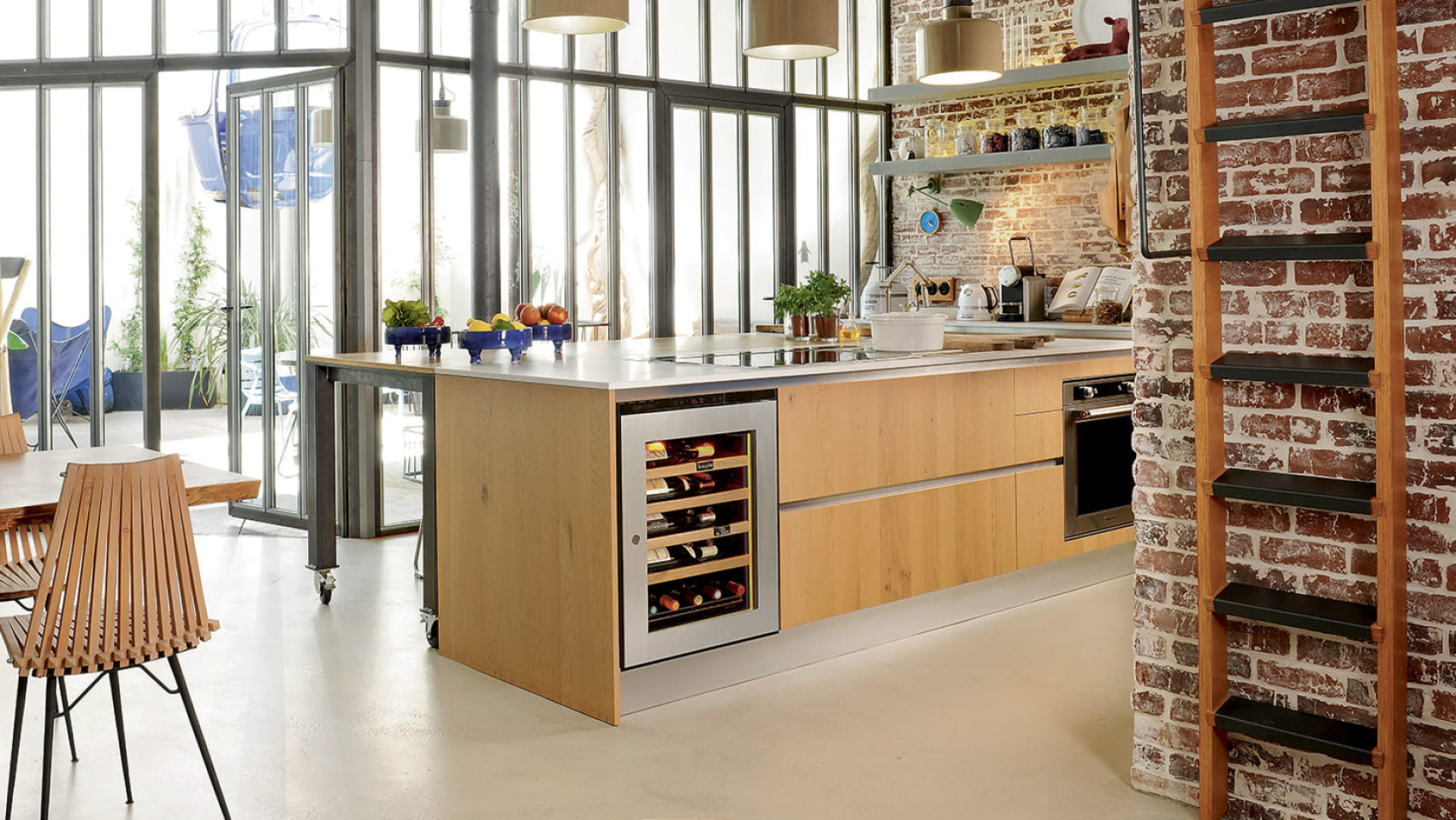 Kleiner Weinklimaschrank mit Glastür und Edelstahlrahmen in einer offenen Küche mit Erkerfenster im Industriedesign, Backsteinmauer und hellen Holzmöbeln.